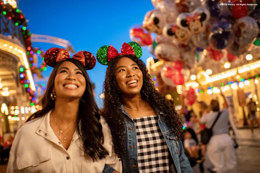 Two young women with sparkly green and red mickey ear headbands on stood on main street. The buildings in the background are lit and there are balloons in the background.