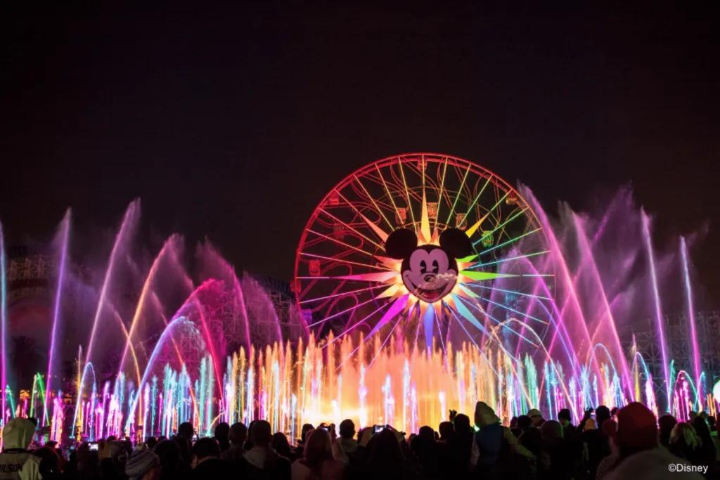 Huge ferris wheel lit up in the different colours of the rainbow with huge Mickey Mouse's face smiling in the middle. In front there are huge fountains which look like they're almost as tall as the Ferris wheel lit up pink. There are so many. 