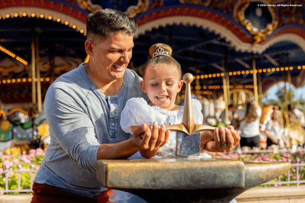 Dad helping his daughter who is dressed in a blue princess dress, pull out a sword from a stone. They are stood in front of the carousel.