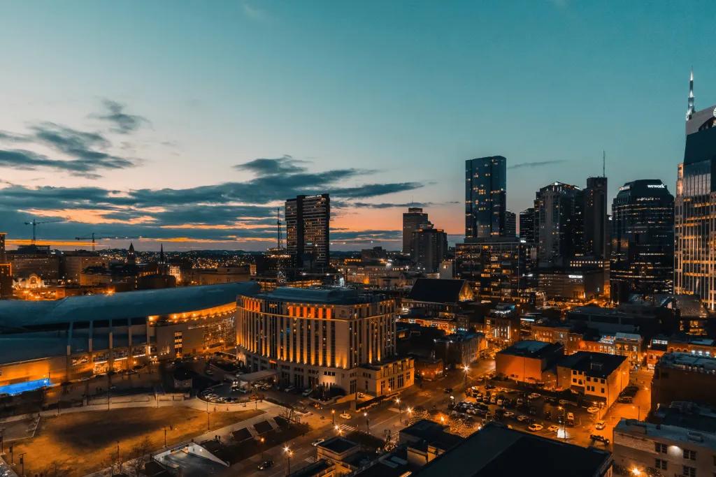 A city at sunset with all of its lights o. There are many sky scrapers and a few smaller buildings. The lights are an amber colour matching the sky.