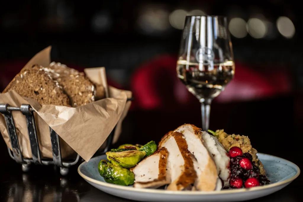 A bread basket next to a plate of chicken, cranberry sauce, Brussel sprouts and other vegetables. Behind there's a glass of wine.