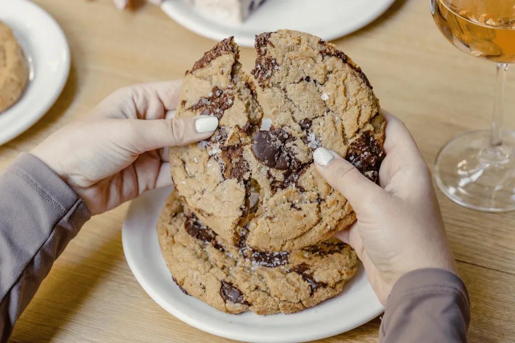 Two huge delicious cookies, one on a white plate and the other in a woman's hands. She is holding it either side a breaking it in half to show the chocolate chip inside.
