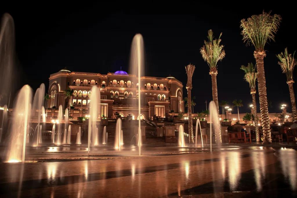 A grand hotel which looks big enough to be a palace with fountains in front, some about 10 foot tall and one in the centre approximately 20 feet tall. The building is lit in the night time a golden colour, as are the fountains. There is a small dome on the roof which is coloured blue.