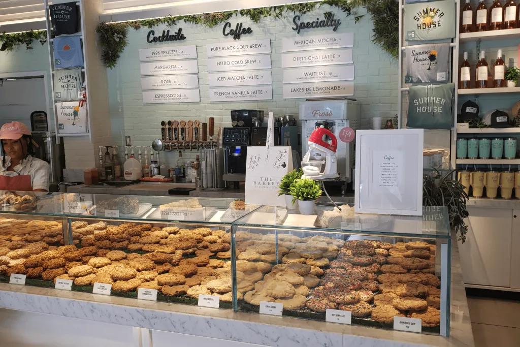 A bakery counter with an array of different huge cookies, a mixer machine on top and behind the counter there's loads of menus showing the different food and drink options.