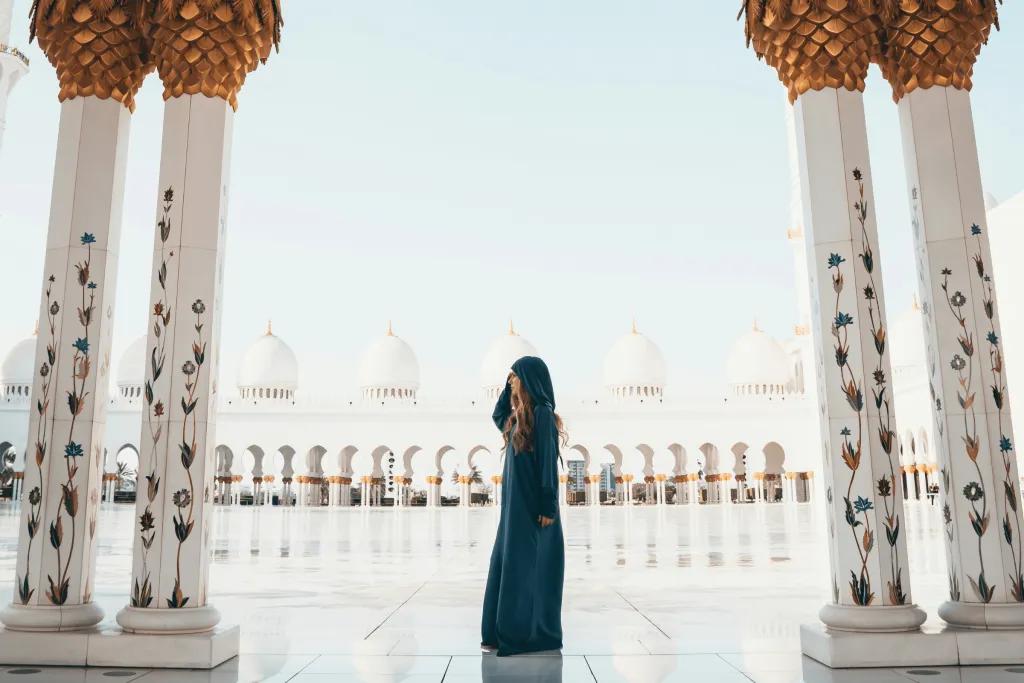 There is a gorgeous sleek tiled floor which is so clean you can see a reflection of people in it. There's a woman wearing a robe which covers her whole body to be respectful to the culture. There are pillars which are white and gold with paintings of flowers growing up them and on the other side it is the same, with white domes on top of the pillars.