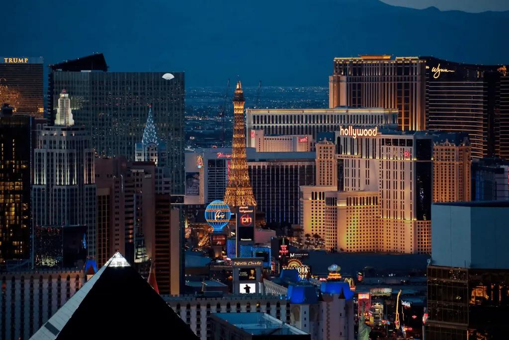 The strip of Las Vegas. There is a straight road with cars on it and a large pointy metal building coloured yellow with lighting, a replica of the Eiffel Tower. There is also a huge air balloon lighted in neon blue and yellow. There are many casinos and hotels as well.