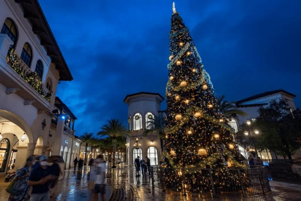 Huge christmas tree outside in the rain with large gold baubles, tinsel and a star on top. Around, there are white buildings surrounding with shops in them. 