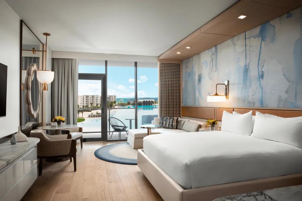An immaculately clean hotel room with wooden, white and blue colouring. There are low hanging lights on either side of the room and a clean white sheet bed. The window overlooks the huge pool outside.