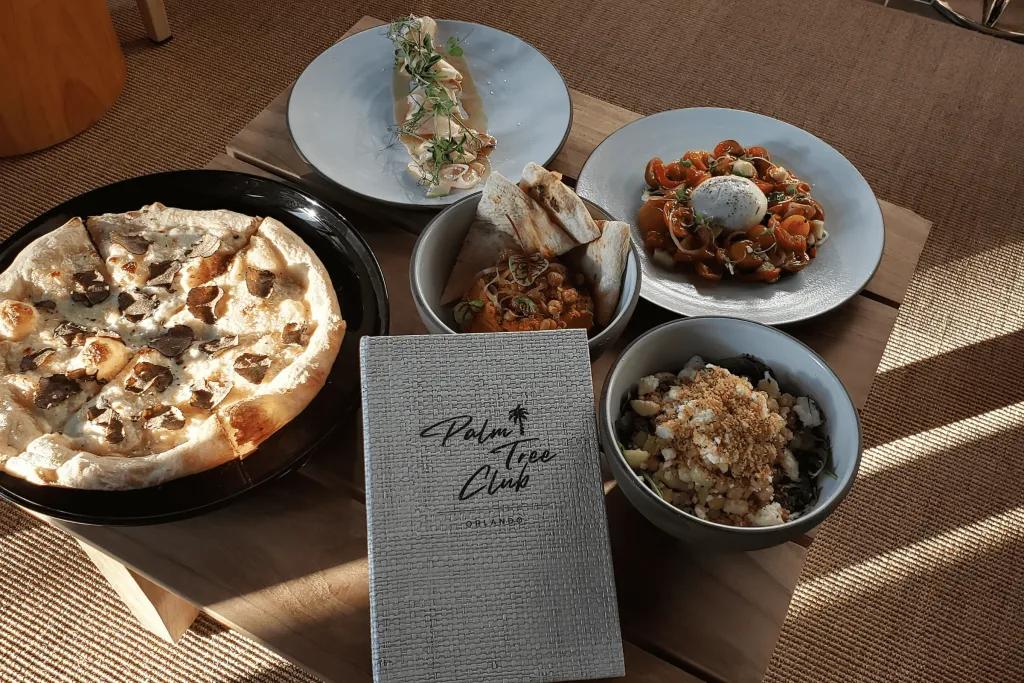 There is a grey menu saying the Palm Tree Club on it and surrounding this there are 5 different dishes. One garlic bread, one mac and cheese and a few other delicious dishes.