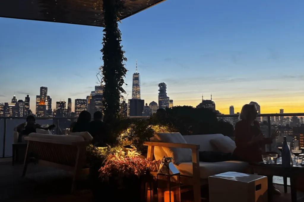 Rooftop bar in new york city. The sky is a dark blue with a hint of yellow and orange as the sun sets in the evening. There are lamps on the ground with candles in them, flowers next to it and plants weaving around the wooden bar beams. This bar is outside  and people are sat talking on sofas and the view shows the city skyline lit up.