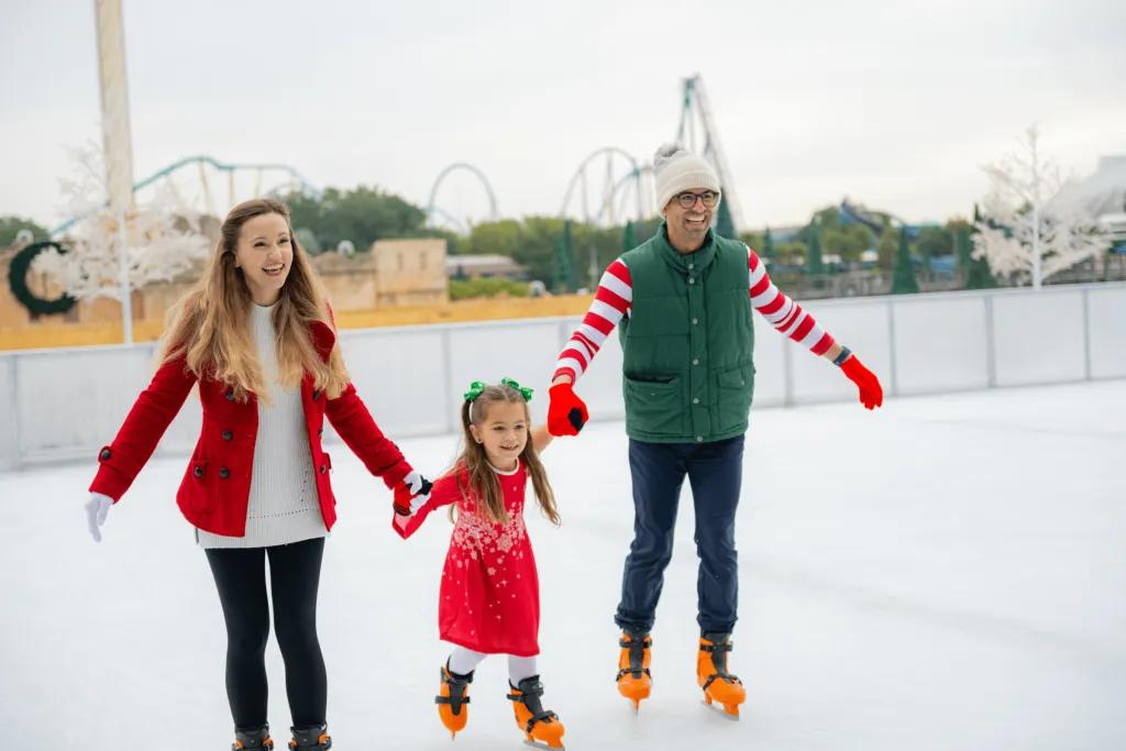 Two parents holding their little girls hand on an ice rink, with the little girl skating in the middle. They are all smiling and in the background you can see the tall rides.