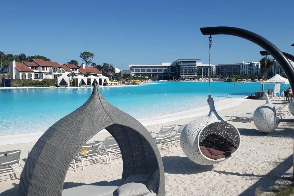 Tear drop shaped hammocks and sun beds made to create shade for guests. They are different shades of grey. A couple are hanging. They are on white sand with white deck chairs behind overlooking the beautiful crystal clear swimming pool.