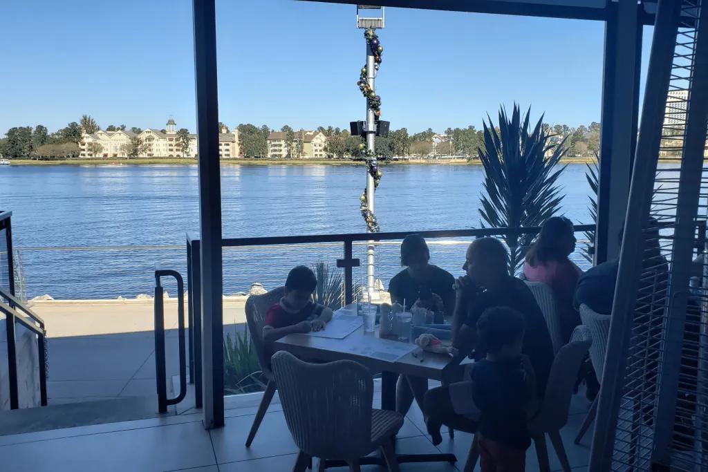 A beautiful terrace with people dining at tables overlooking a large lake. There are posts with Christmas decorations swirling up them. On the other side of the lake there are pretty white buildings.