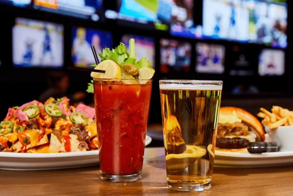 A large bloody mary with celery on top and a large beer in front of delicious food. There is a huge plate of loaded nachos and on the other plate there is a burger with a side of fried. In the background, you can see many tv screens with different sports playing.
