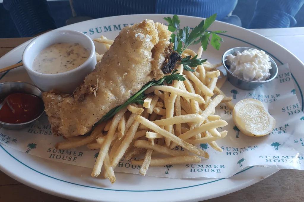 There's a plate with the name of the restaurant written on it. There is a delicious serving of fish and chips with pots of sauces next to it.