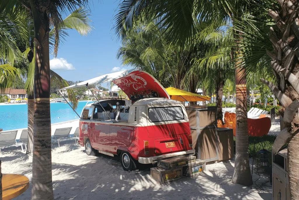 On the white sand back there is a red campervan renovated into a bar. In the roof which is lifted up, there are bottles of alcohol. There are many palm trees surrounding this bar and the pool is in the background.