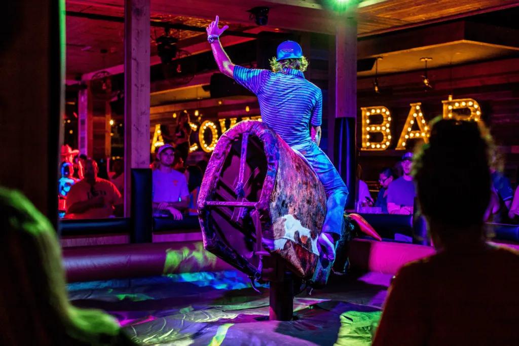 Man with backwards cap on is on a mechanical fake bull, holding on tight and trying to stay on as it spins around and everyone is crowded watching around the sides of the black inflatable. There are beams everywhere and in the background you can see "A COWBOY BAR" in large lights on the wall.