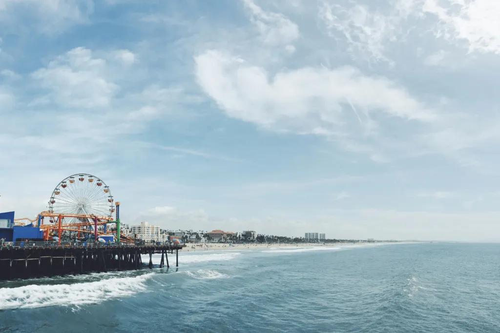 There is the sea waves crashing onto a yellow sandy beach. There is also a long iconic pier with a funfair on it, including a Ferris wheel and small rollercoaster. In the far distance there are only a few large buildings..