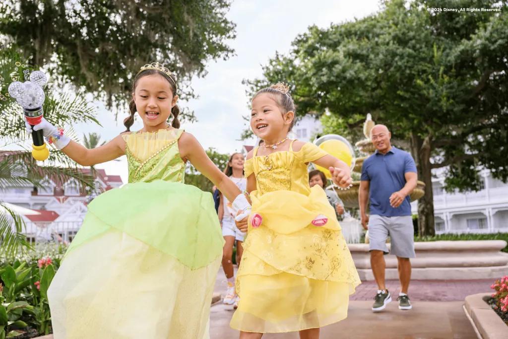 Two young girls running through a resort in Walt Disney World Resort. The girls are dressed as Tiana and Belle with dresses on.