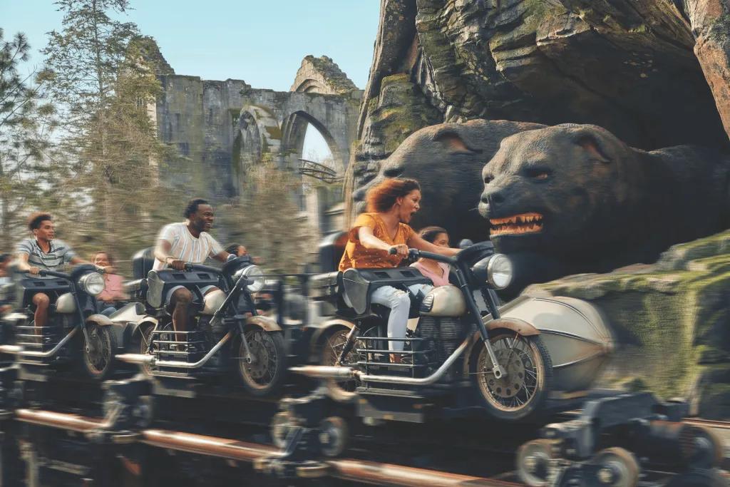 Guests on motorbike shaped vehicles and sidecars zooming past a three faced giant dogpoking out of a cave and there are ruins in the background.