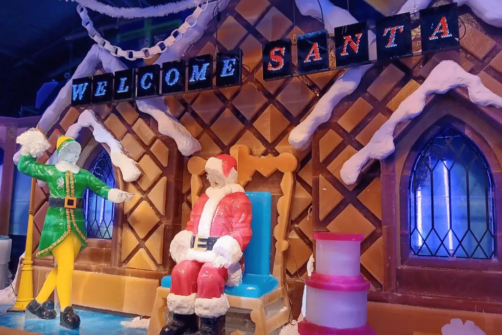 Ice sculpture of santa sat on a throne in front of a gingerbread house. Above the gingerbread house, there are blue and red signs saying "welcome Santa" and to the left, there is a buddy the elf  sculpture throwing a lump of snow at Santa.