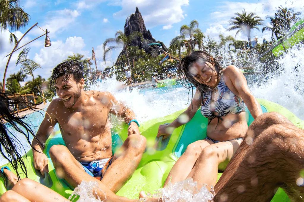 4 people sat in a big rubber ring heading into a large pool of water which is splashing them as they cling on. In the background there is a large brown/grey coloured volcano.