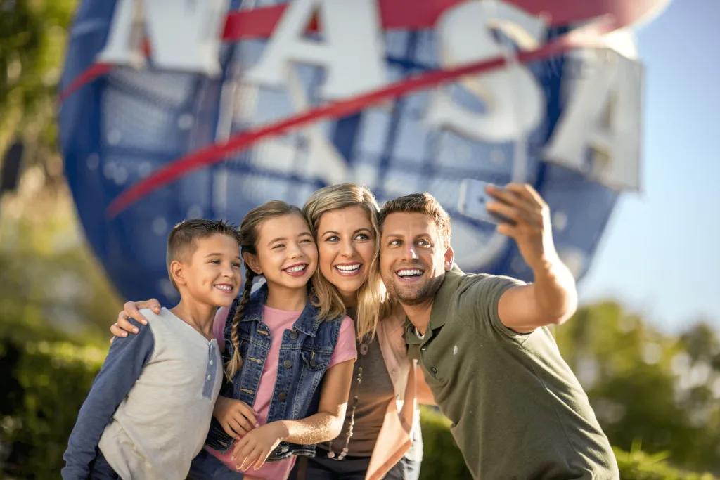 Family of four smiling brightly, 2 kids and 2 parents taking a selfie in front of a large blue globe statue with red lines zooming across and the word NASA.