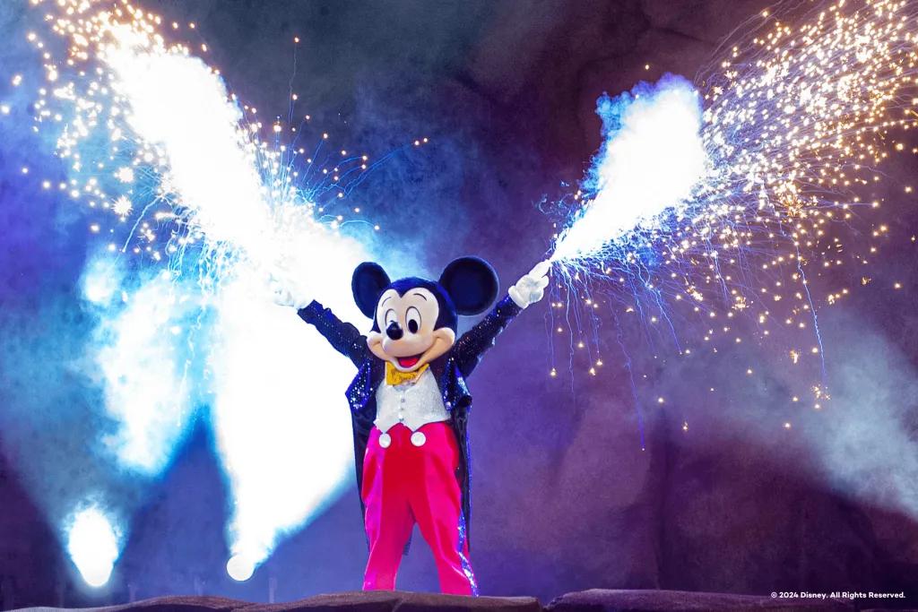 Mouse with black ears and beige face wearing a long black coat, red trousers and a white shirt. He has sparklers coming out of both of his hands.
