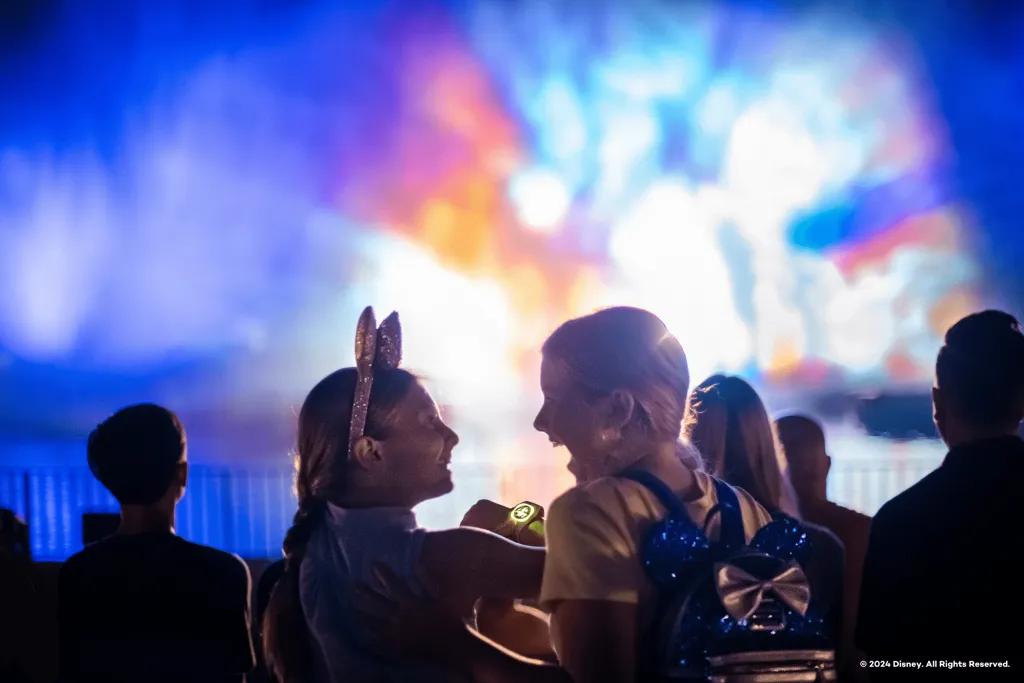 Two little girls smiling and gasping looking at one another and in front of them are giant fountains with Disney movies projected onto them