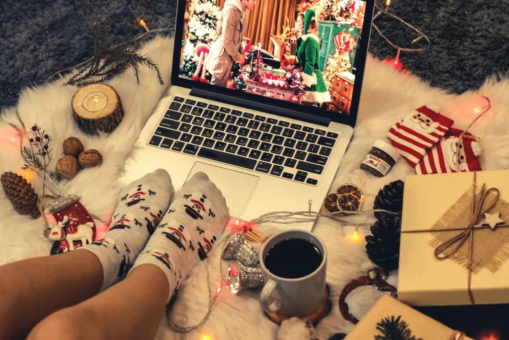 Someone sat on a white blanket with christmas lights, watching a christmas movie on their laptop with decorations around like stripey socks and presents
