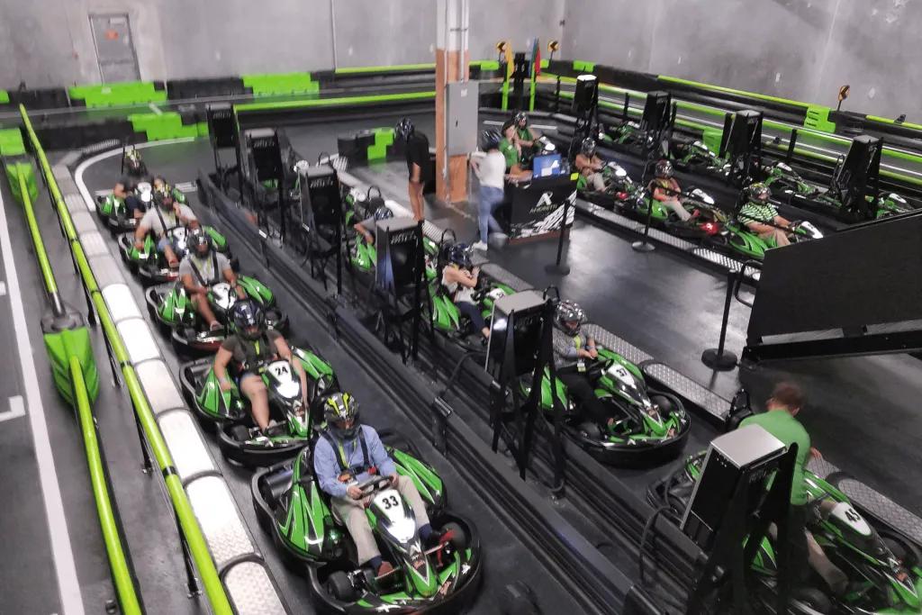 Black and green go karts with people sat in them waiting to start their race. There is a black track and they are all wearing helmets.