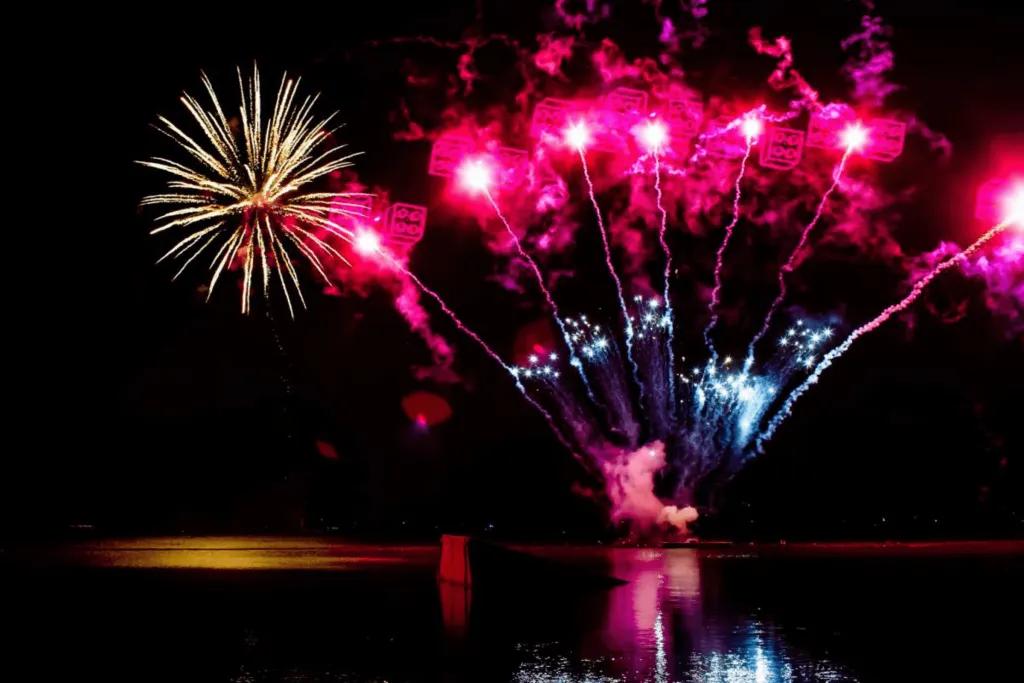 Red fireworks bursting over lake at night with red LEGO blocks in the sky. There is also a huge gold firework in the sky.