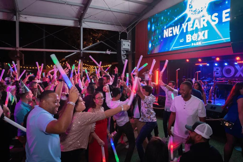 Crowd of people holding up light sticks and dancing looking towards a screen which says New Years Eve Boxi and a sparkling disco ball.