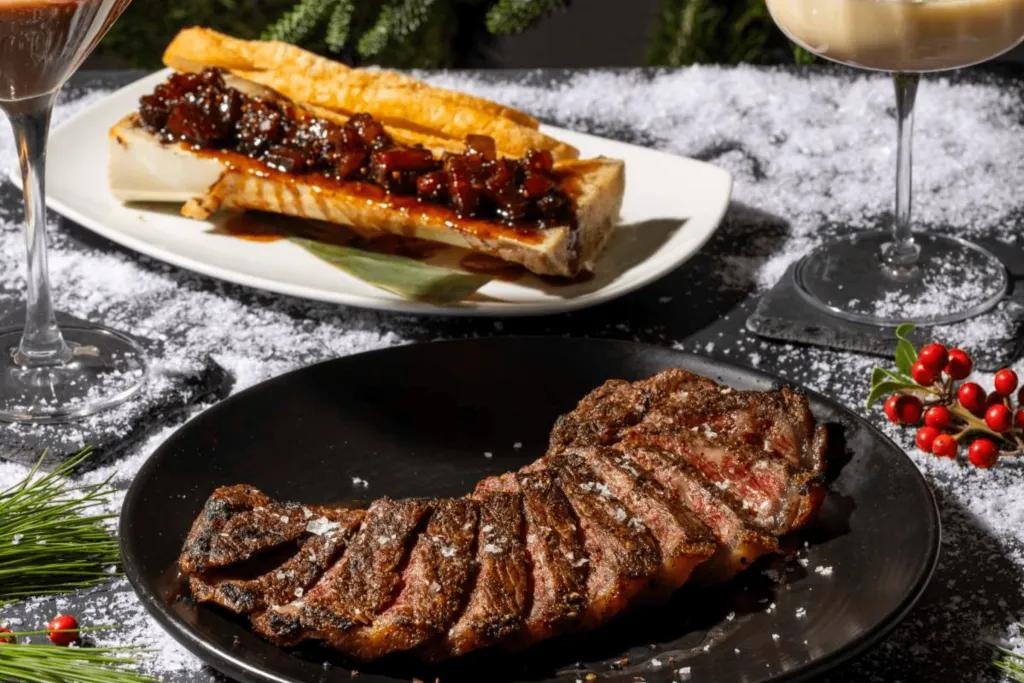 A black plate with steak cooked medium well, some bread with a sauce on it and a glass of wine on the side.