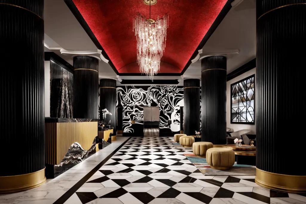 Dark lounge with a patterned black and white tiled floor has a red ceiling and a chandelier hanging down. There are beige chairs for people to lounge on. 