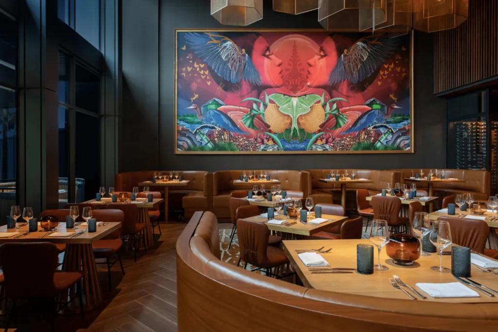 A large restaurant with huge painting on the wall which includes two red people symmetrically looking at one another. The tables are light wood and dark chairs.