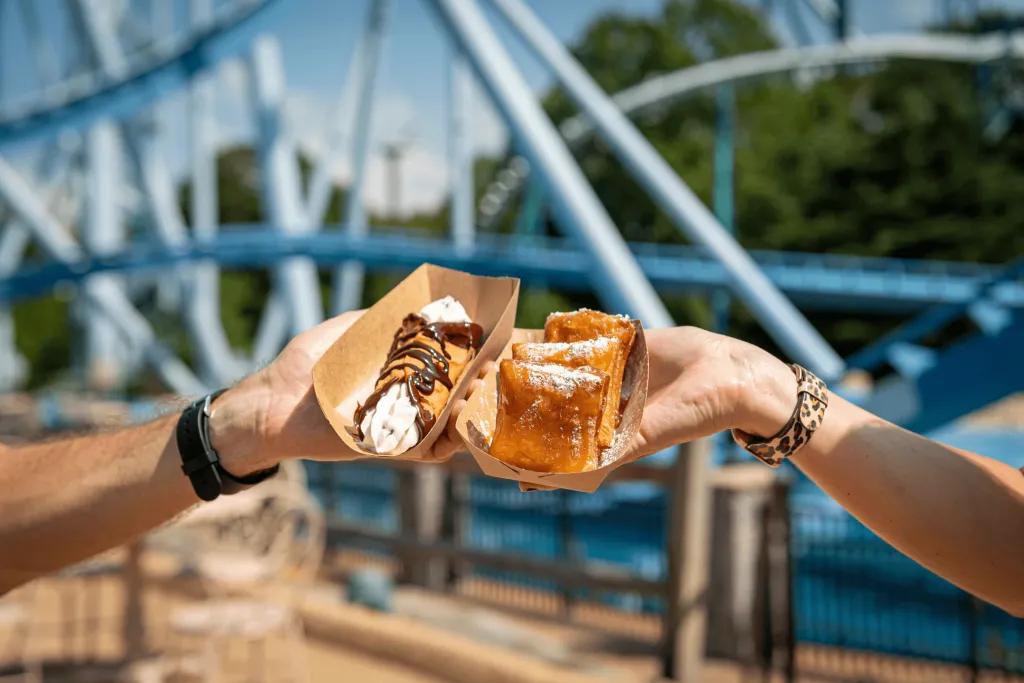Two hands holding two dishes in front of a rollercoaster track. One is beignets and the other is a crepe with whipped cream on.
