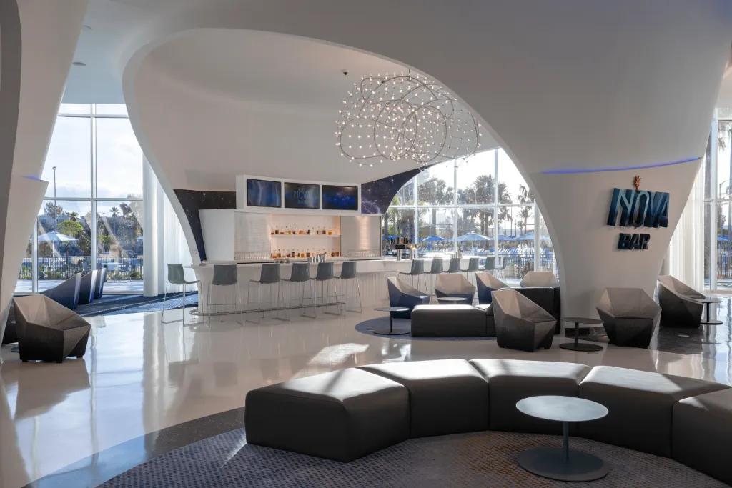 Modern sleek design of lobby with crystal chandelier hanging. There is a bar with stalls and brown sofas around small tables.