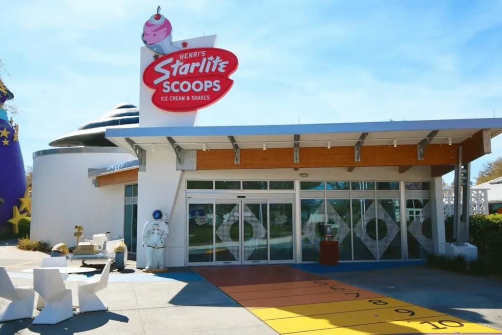 A diner styles modern building with white walls and window panels around. There is a tall sign with white writing saying Henri’s Starlite Scoops, and an image of a giant ice cream.