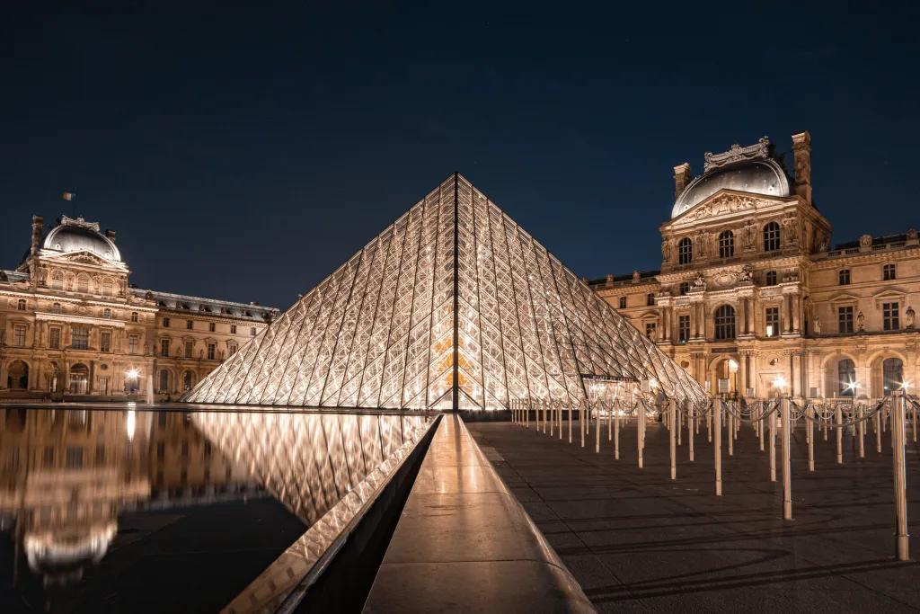 A glass panelled pyramid building in the centre of old fashioned castle like buildings. It is reflecting in the small pond.
