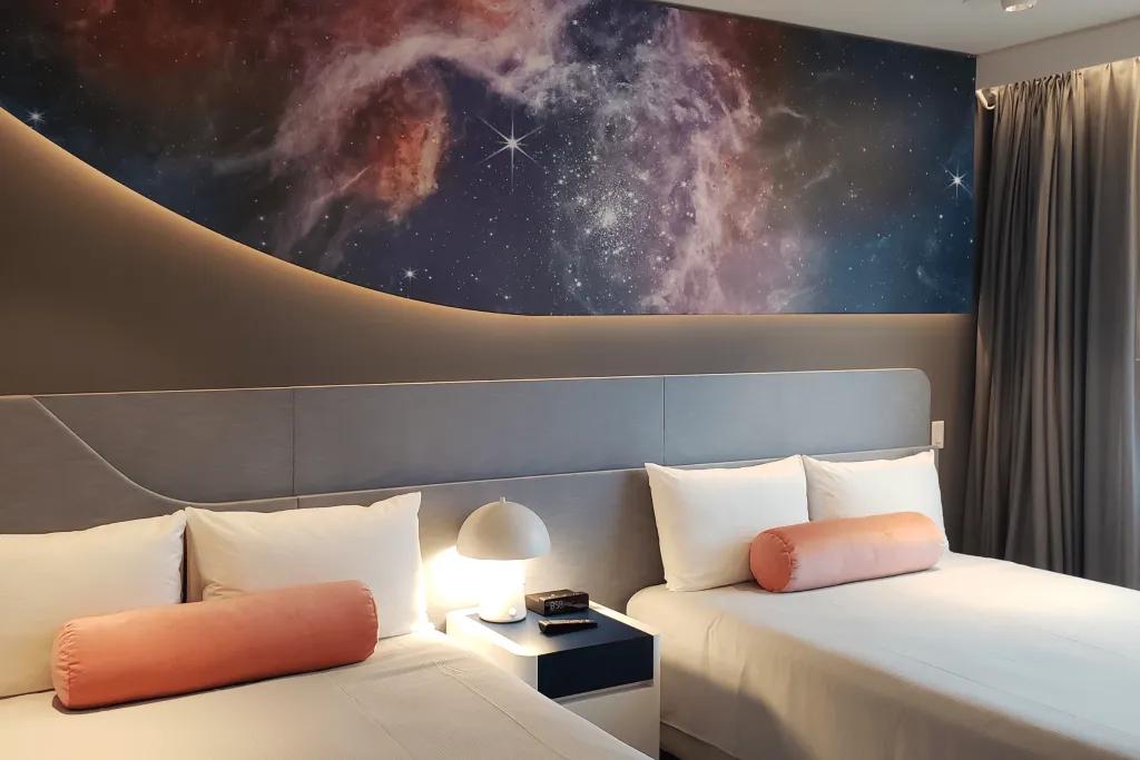 Room with two white beds and orange cushions. There is a small white bedside table with a lamp and above on the dashboard there's images of stars in the universe.