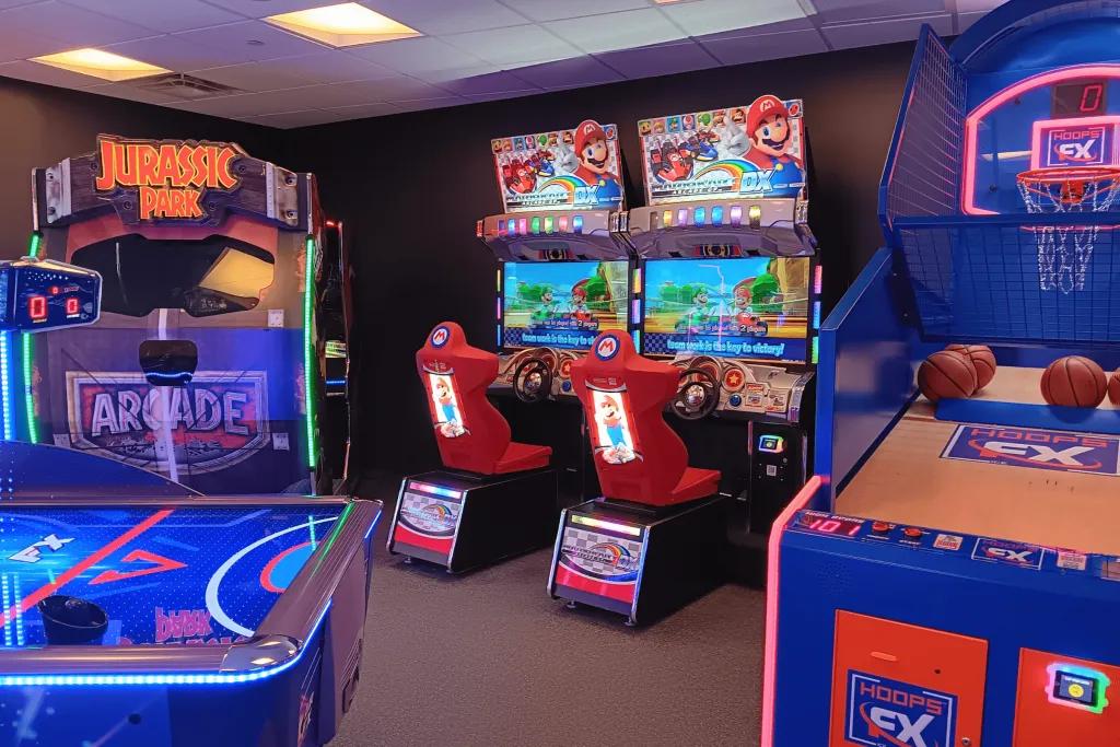 Arcade room with air hockey machine, basketball machine, mario kart driving machine and jurassic park arcade game.