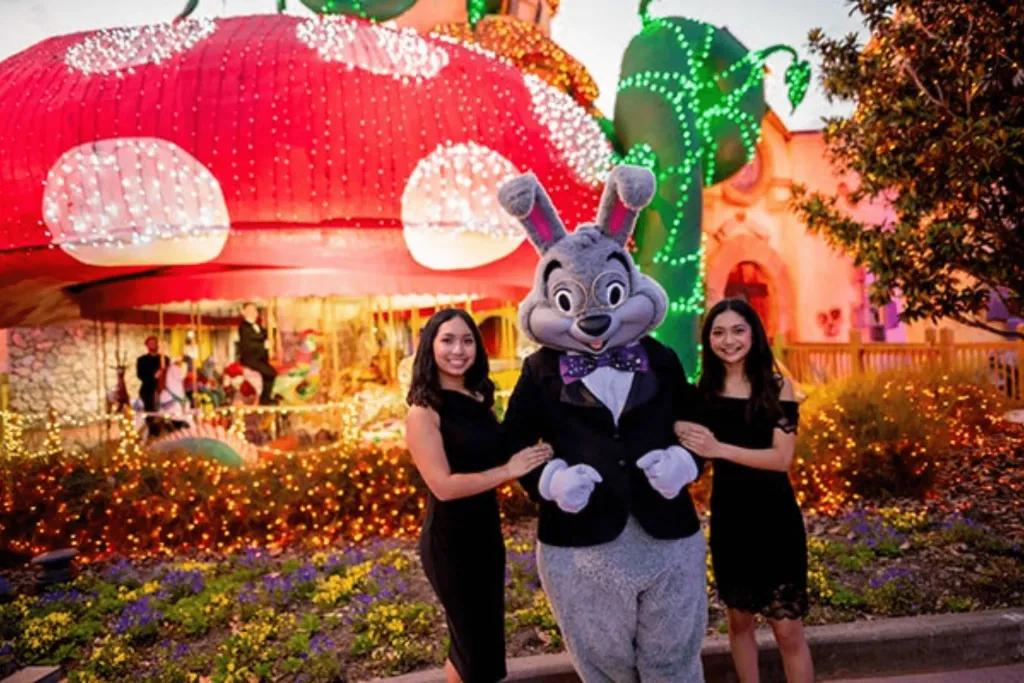 A bunny dressed in suit stood with two young girls in front of a giant toad stall with a carousel underneath.