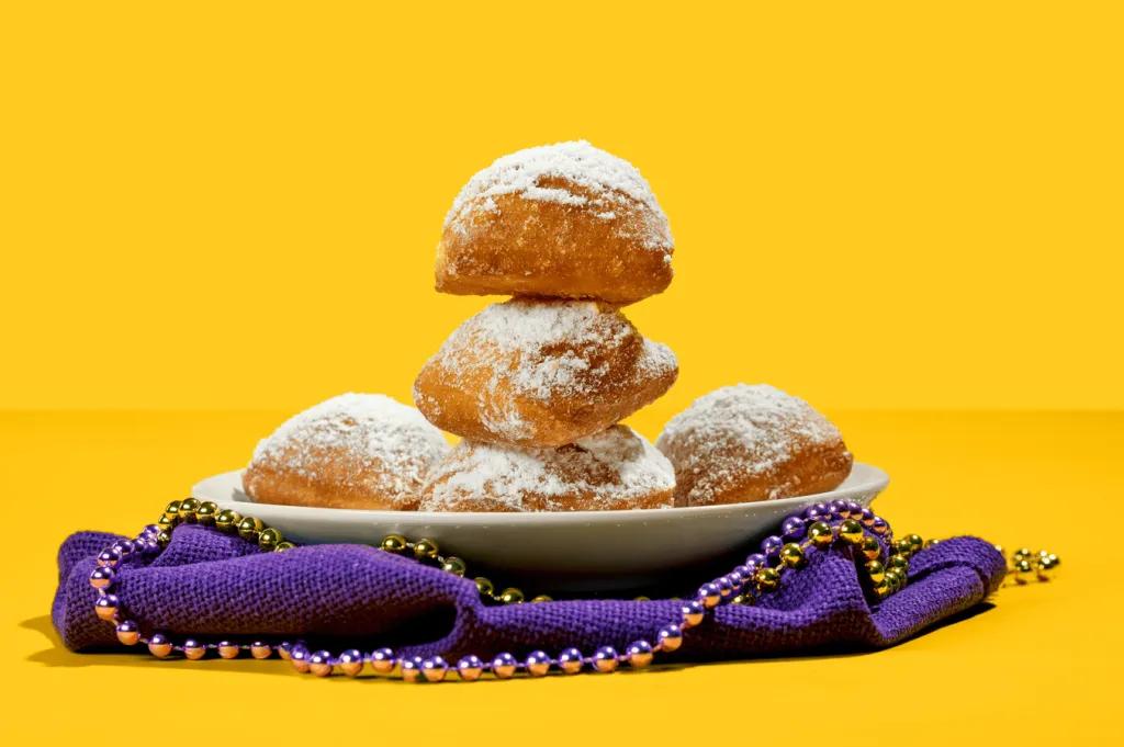 A stack of beignets on a plate on top of a purple napkin and Mardi Gras beads