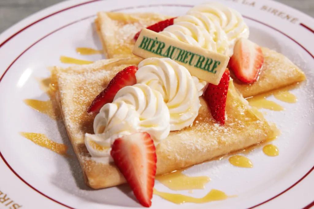 Two crepes folded into quarters with whipped cream on, yellow sauce drizzled over and strawberry slices.