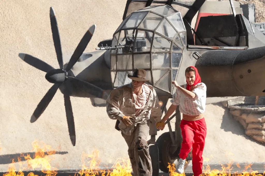 Indiana Jones, a man dressed in explorer clothes and his sidekick, a woman wearing old fashioned clothes, running from a burning aeroplane about to explode, holding each others hands.