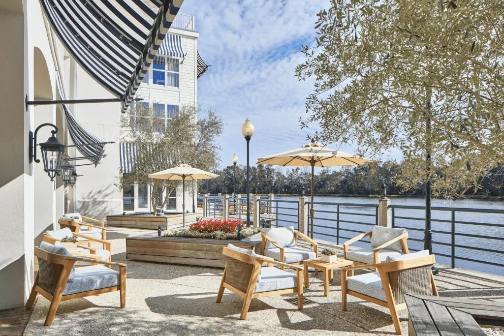 Outside the large white hotel on the edge of a large lake in the sun, there are clean cream decking chairs, tables and umbrellas. 