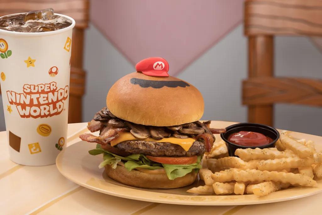 A large burger with: a brioche bun, large beef patty, cheese, mushrooms, lettuce, tomato and a little mario red hat on top. There are fries and a small black pot of ketchup. There is also a large cup of coke.