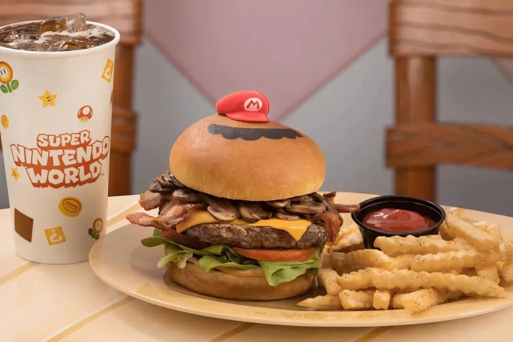 Ein großer Burger mit einem Brioche-Brötchen, einem großen Rindpatty, Käse, Champignons, Salat, Tomate und einer kleinen roten Mario-Mütze darauf. Dazu gibt es Pommes und einen kleinen schwarzen Topf mit Ketchup. Außerdem gibt es einen großen Becher Cola.