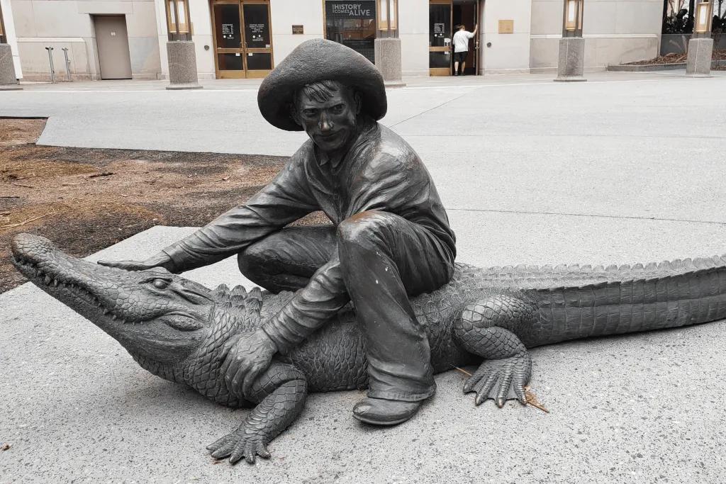 Statue of man sat on alligator holding its head and arm, wearing a large hat.